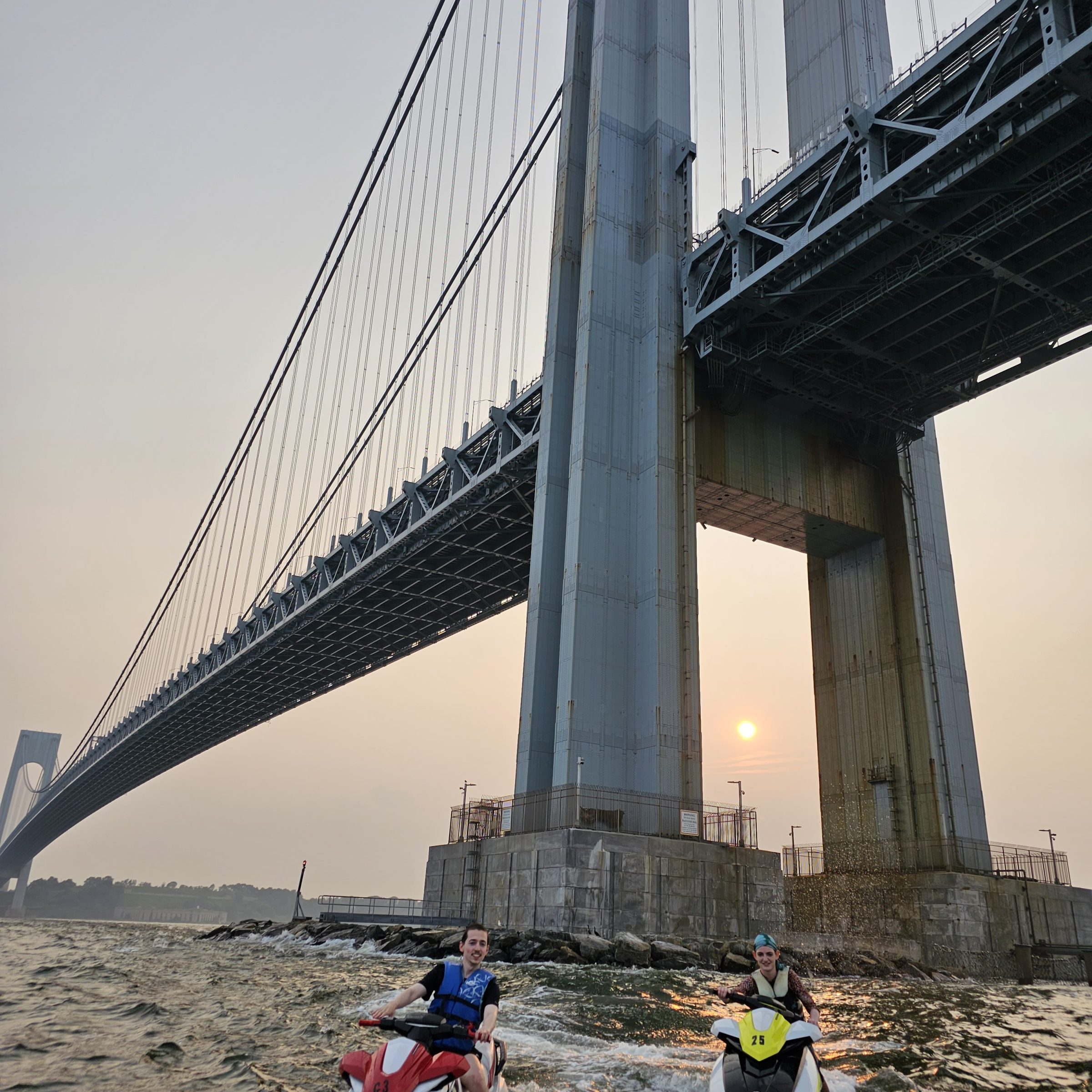 People on jet skis near a large suspension bridge at sunset.