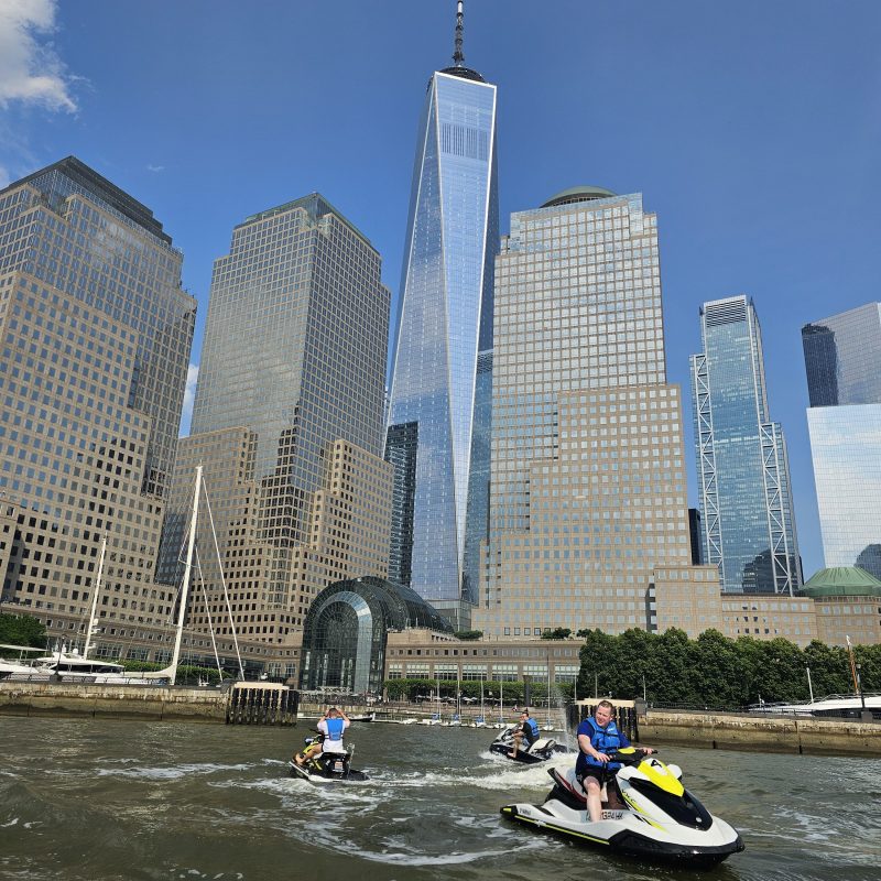 People on jet skis in river near tall city buildings, under clear blue sky.