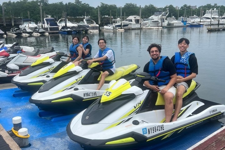 Five people sitting on jet skis at a marina with boats in the background.