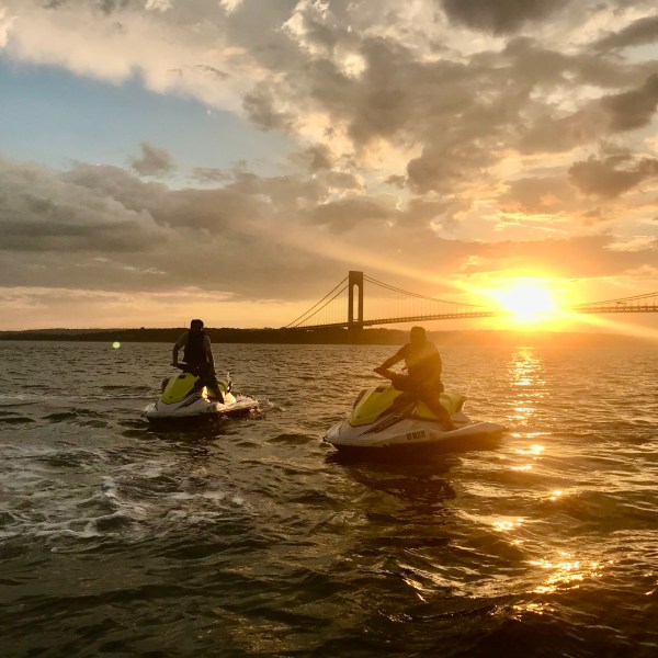 Two people on jet skis in water at sunset near a bridge.