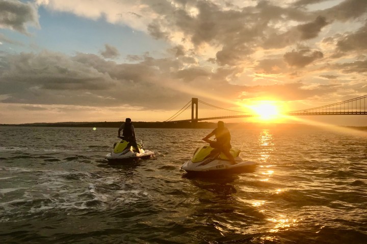 Two people on jet skis in water at sunset near a bridge.