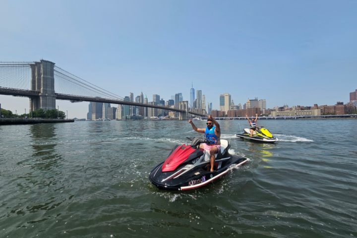 Two people on jet skis in a river with a city skyline and bridge in the background.
