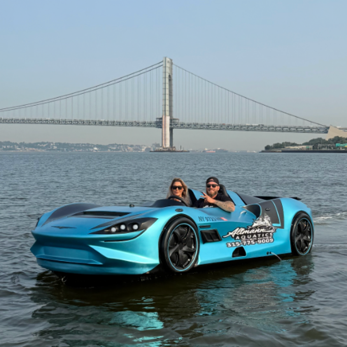 Two people in a blue sports car-like boat on water with a bridge in the background.