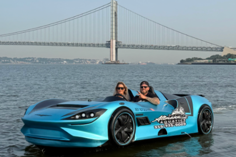 Two people in a blue sports car-like boat on water with a bridge in the background.
