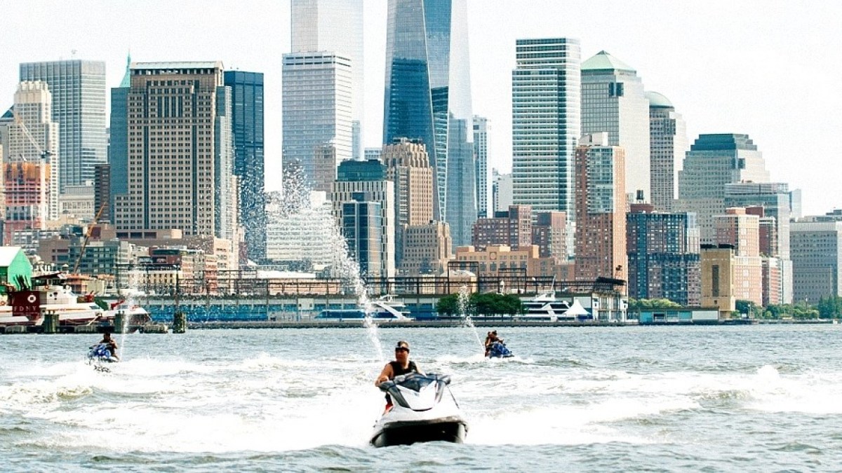 People on jet skis ride in river with city skyline in background.