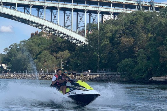 Person riding a jet ski under a bridge on a sunny day.