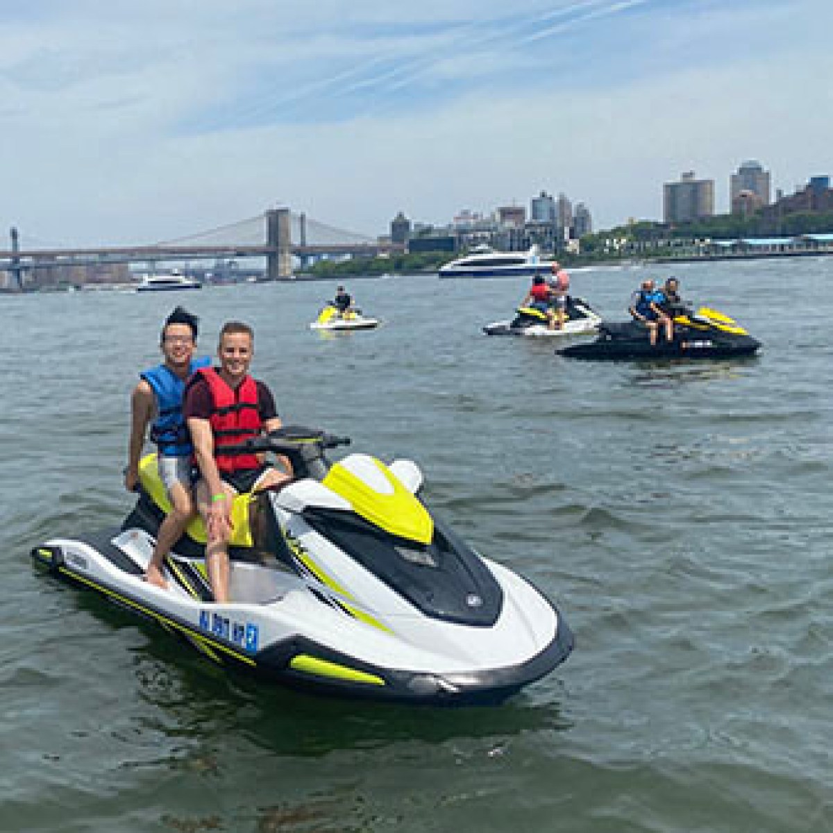 People riding jet skis on a river with a city skyline in the background.