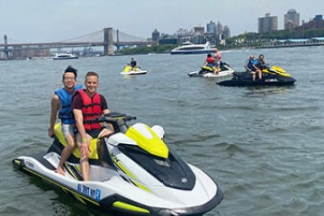 People riding jet skis on a river with a city skyline in the background.