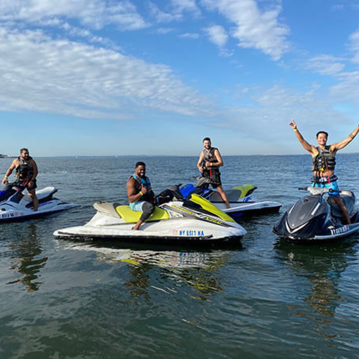 Four people on jet skis in a sunny lake, one with arms raised.