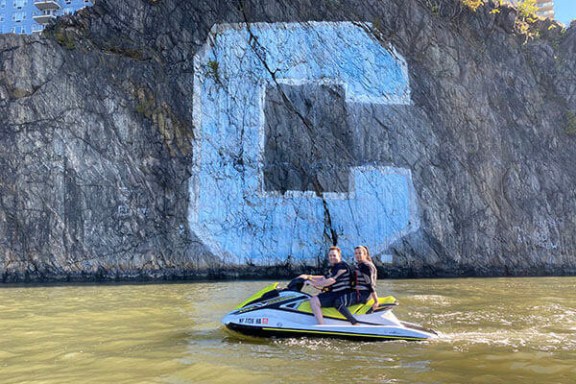Two people on a jet ski near a rocky cliff with a large blue 'C' painted on it.