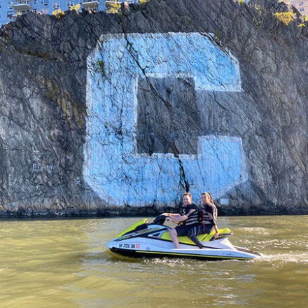 Two people on a jet ski near a rocky cliff with a large blue 'C' painted on it.