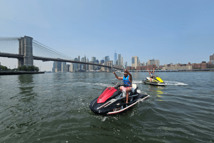 Two people on jet skis in water with city skyline and bridge in background.