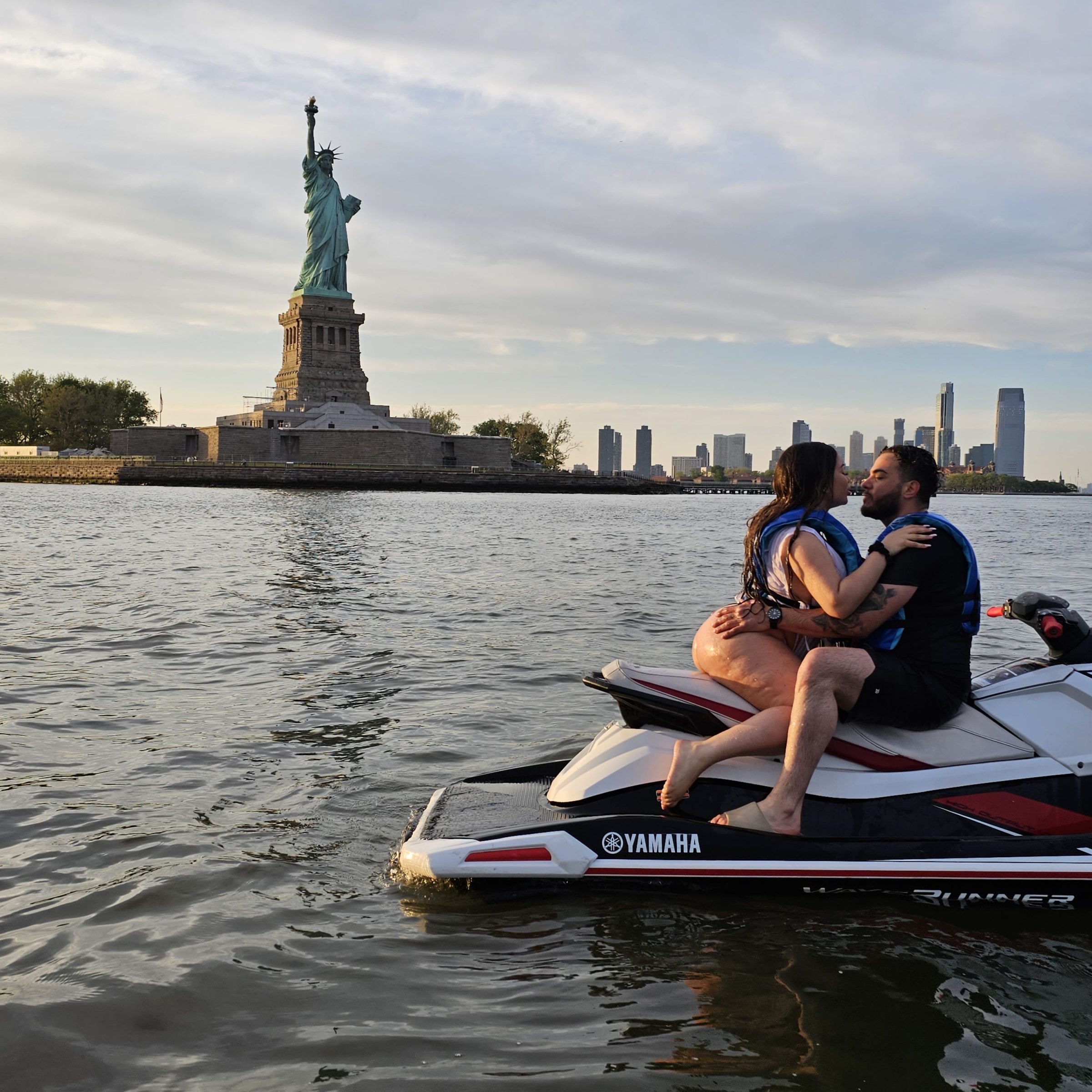 Two people on a jet ski near the Statue of Liberty, city skyline in the background.